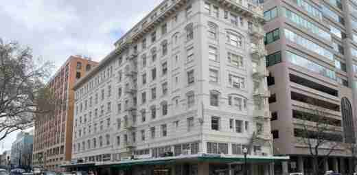 A street view of a historic multi-story white building with ornate architectural details stands surrounded by other buildings. Nearby, a crane from a local crane rental company adds modern contrast as it assists in construction, while trees line the street and cars are parked along the curb.