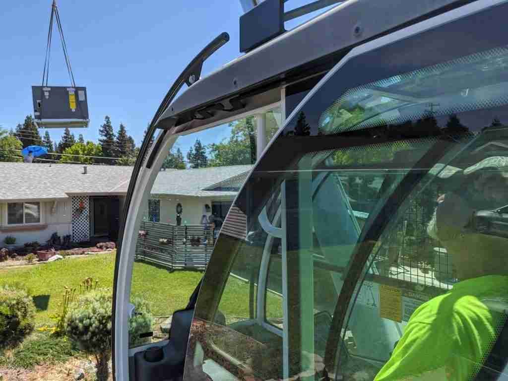 A person in a bright green shirt operates a mobile crane from inside a cab, lifting a large HVAC unit above a residential lawn. A white house, trees, and a clear blue sky are in the background, showcasing the versatility of crane rentals in suburban settings.