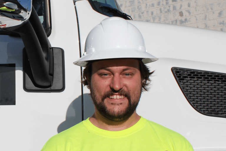 A person wearing a white hard hat and a bright yellow shirt stands in front of a white truck, symbolizing the efficiency of crane rentals. They have a beard and are smiling at the camera, with part of a brick wall visible in the background.