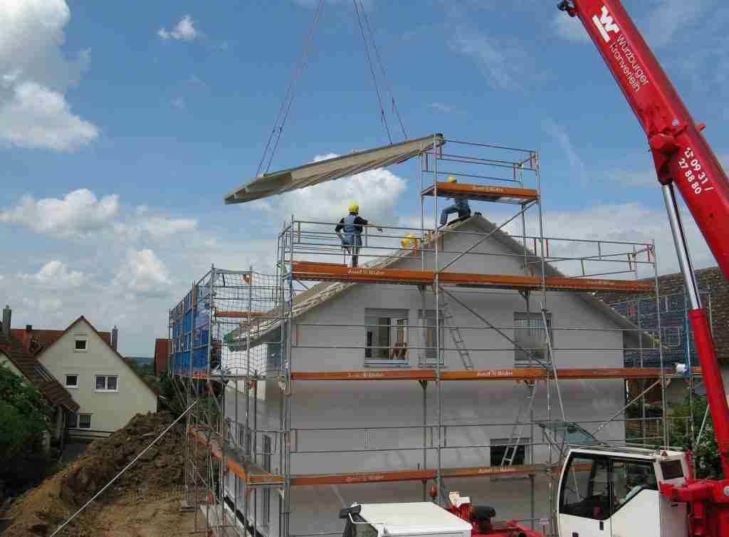 A house under construction is encircled by scaffolding. Two workers on the roof guide a large panel being lifted by a red mobile crane from a renowned crane company. The sky is blue with sparse clouds.