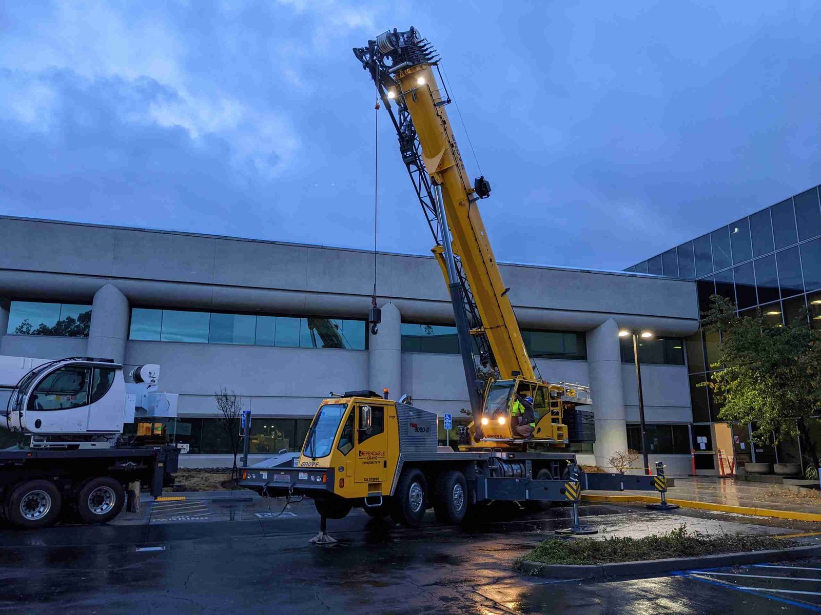 A large yellow mobile crane is positioned in a wet parking lot at dusk, with its arm extended. Nearby, a building with large windows reflects the evening sky. The area, dimly lit, exudes a calm, industrial atmosphere perfectly suited for crane rental needs.