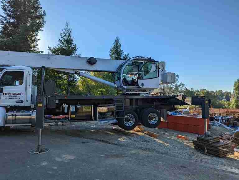 A crane truck with "Dependable Crane" written on it is parked on a construction site, showcasing the reliability of our crane rentals. The crane's arm is extended, and the truck is stabilized on outriggers. Construction materials and trees are visible in the background under clear skies.