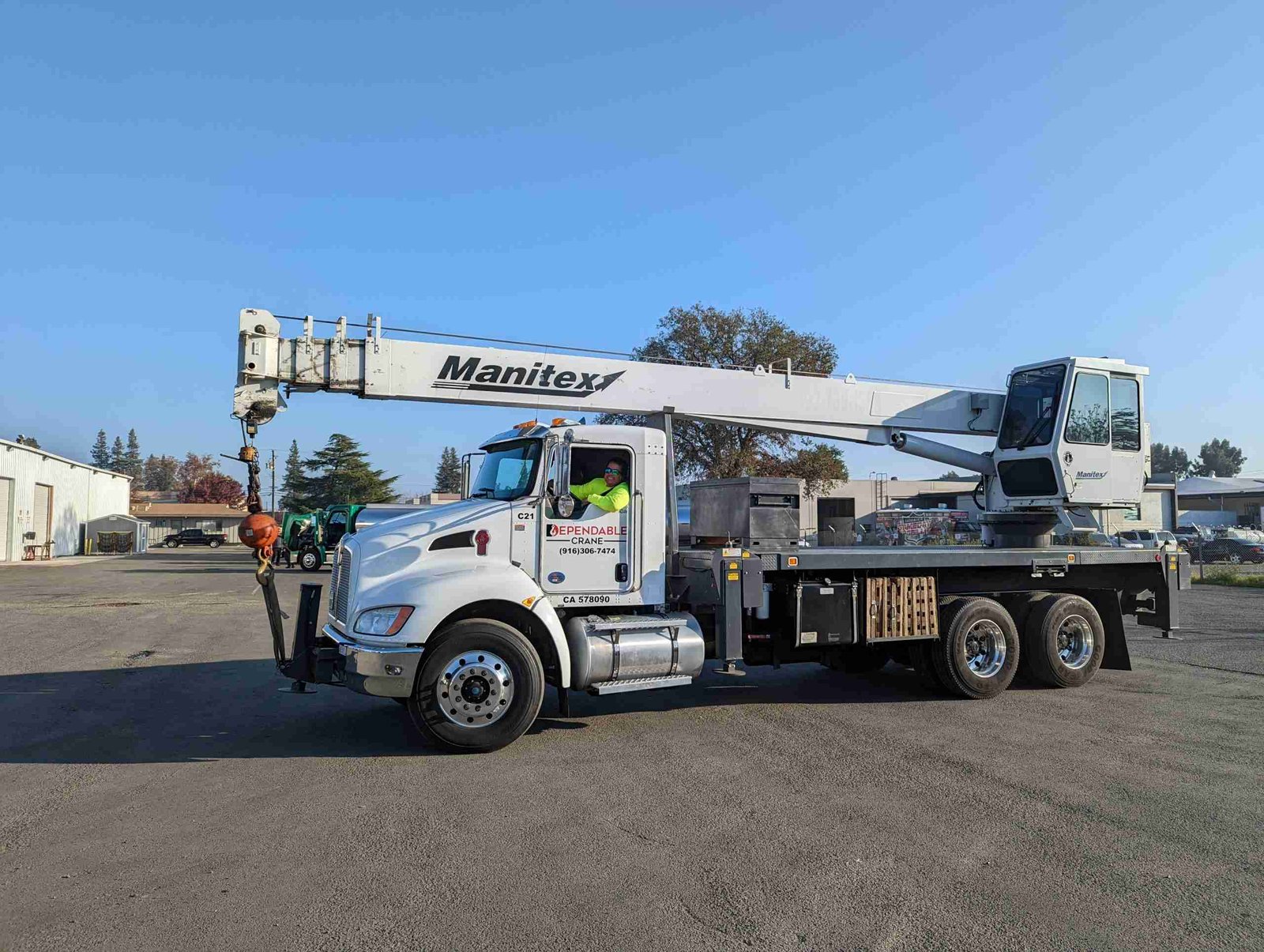 A truck with a Manitex crane, available for crane rentals, is parked on an asphalt lot under a clear blue sky. The crane's arm extends over the truck, and a worker in a safety vest is visible in the cabin. Nearby are industrial buildings and some distant trees.