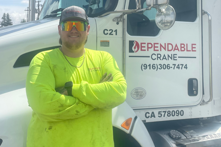 A person in a bright yellow shirt and cap stands smiling with arms crossed in front of a white truck. The truck, showcasing "Dependable Crane," highlights their crane rental services, with contact details prominently displayed on the side.