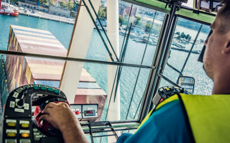 A crane operator in a yellow safety vest expertly manages crane operations, maneuvering levers inside a cabin while overseeing the bustling shipping port. Numerous stacked shipping containers are visible on the vessel below, underscoring the crucial role of crane safety in this dynamic environment.