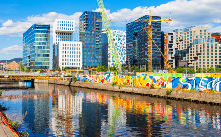 A vibrant urban waterfront features a row of modern, uniquely designed buildings under a blue sky. A crane, part of innovative crane solutions, is visible amidst construction. The water reflects colorful street art on a wall along the canal, adding dynamic color to the scene.