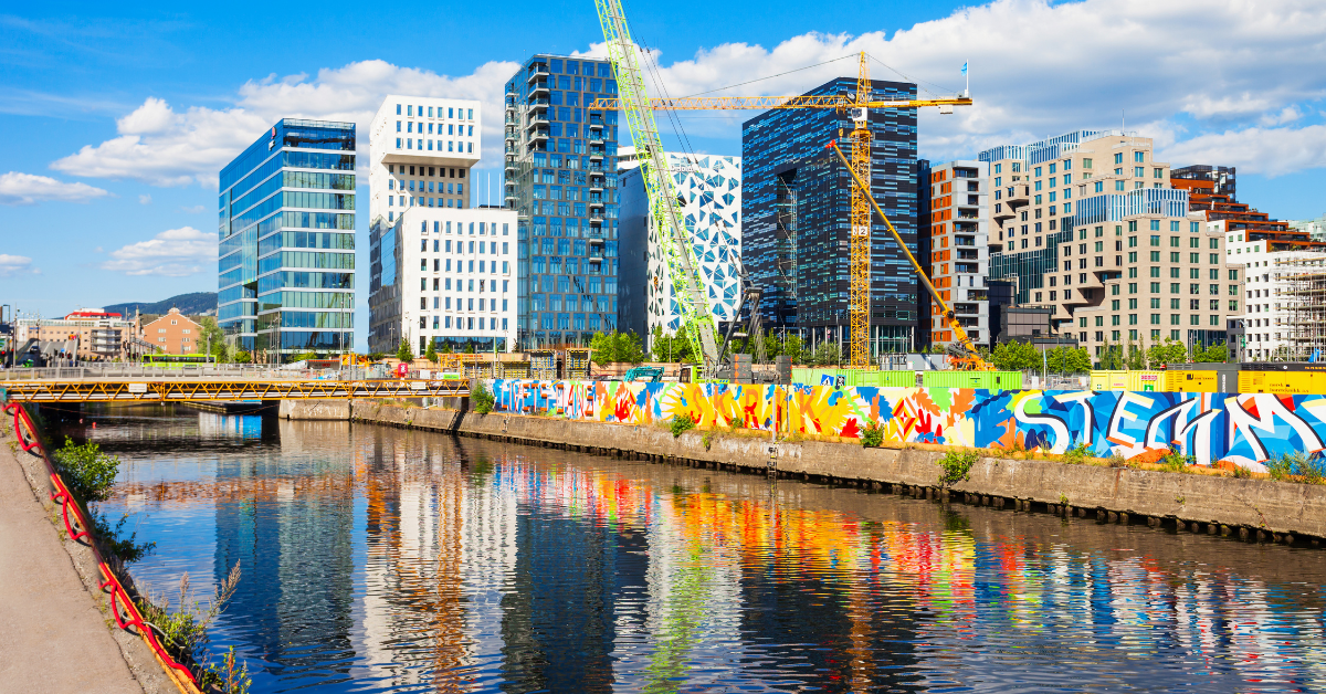 A vibrant urban waterfront features a row of modern, uniquely designed buildings under a blue sky. A crane, part of innovative crane solutions, is visible amidst construction. The water reflects colorful street art on a wall along the canal, adding dynamic color to the scene.