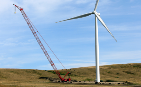 A large red crane is busy with the installation of a tall white wind turbine on a grassy hill. The sky remains clear with a few wispy clouds, enhancing the expansive landscape.