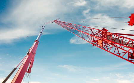 Low-angle view of two cranes set against a clear blue sky with scattered clouds. The cranes' towering structures intersect, one red and one gray, showcasing the power of modern lifting equipment as they create dynamic lines against the backdrop, symbolizing construction and engineering.